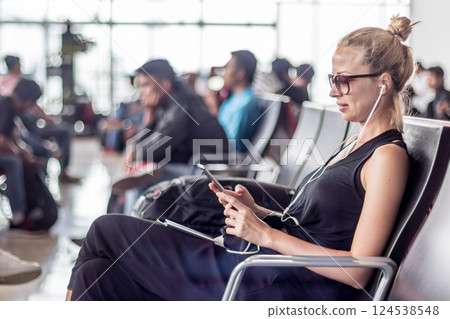 Casual sporty young blond female traveler using her cell phone while waiting to board a plane at the departure gates at the asian airport terminal. 124538548