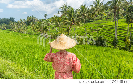 Relaxed fashionable caucasian woman wearing red asian style kimono and traditional asian paddy hat looking at beautiful green rice fields and terraces on Bali island. Relaxed fashionable caucasian woman wearing red asian style kimono and traditional asian paddy hat looking at beautiful green rice fields and terraces on Bali island. 124538551