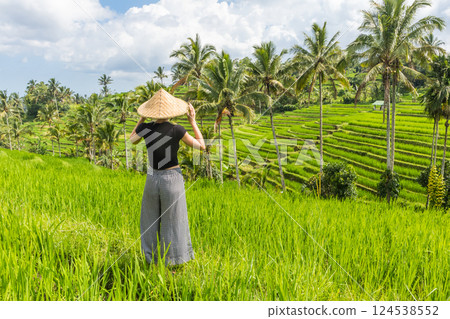 Relaxed fashionable caucasian female tourist wearing small backpack and traditional asian paddy hat looking at beautiful green rice fields and terraces on Bali island. Relaxed fashionable caucasian female tourist wearing small backpack and traditional asian paddy hat looking at beautiful green rice fields and terraces on Bali island. 124538552