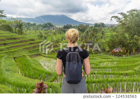 Casual caucasian female tourist wearing small backpack looking at beautiful green rice fields and terraces of Jatiluwih on Bali island, Indonesia. Casual caucasian female tourist wearing small backpack looking at beautiful green rice fields and terraces of Jatiluwih on Bali island, Indonesia. 124538563