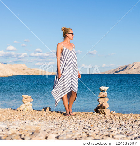 Happy carefree woman wearing beautiful striped summer dress enjoying late afternoon walk on white pabbled beach on Pag island, Croatia. 124538597
