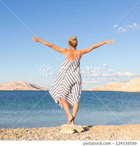 Happy carefree woman rising arms, wearing beautiful striped summer dress enjoying late afternoon on white pabbled beach on Pag island, Croatia. Happy carefree woman rising arms, wearing beautiful striped summer dress enjoying late afternoon on white pabbled beach on Pag island, Croatia. 124538599