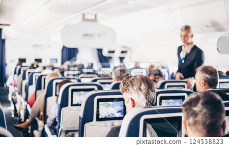 Interior of airplane with passengers on seats and stewardess in uniform walking the aisle, serving people. Commercial economy flight service concept 124538823