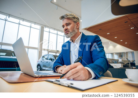 Businessman in suit sitting in automobile dealership office using laptop Businessman in suit sitting in automobile dealership office using laptop 124539355