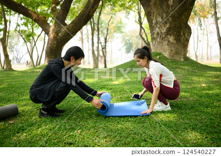 Active couple unroll a yoga mat on lush green lawn, preparing for fitness session together Active couple unroll a yoga mat on lush green lawn, preparing for fitness session together 124540227