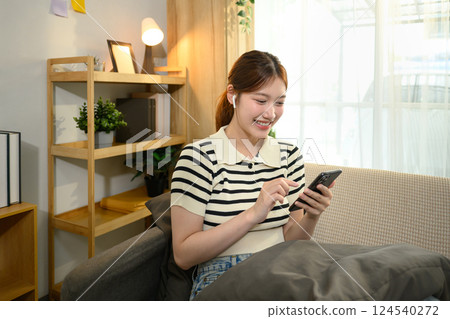 Young woman wearing wireless earphones using smartphone on couch at home 124540272