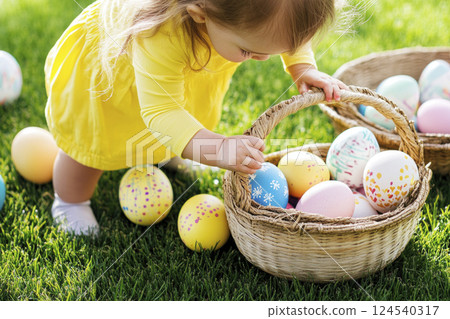 Easter egg hunt. Toddler girl in yellow dress with baskets full of Easter eggs in sunlit meadow. 124540317