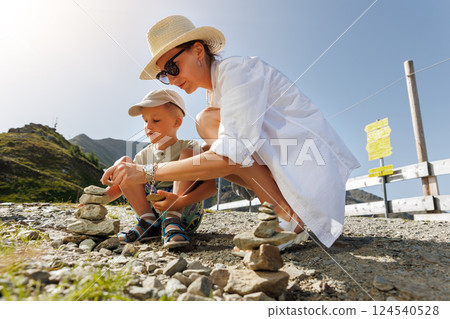 Mother and son build small stone cairns on sunny mountain trail, surrounded by lush scenery. They share bonding moments while enjoying serene alpine beauty and fresh air, forging cherished memories Mother and son build small stone cairns on sunny mountain trail, surrounded by lush scenery. They share bonding moments while enjoying serene alpine beauty and fresh air, forging cherished memories 124540528