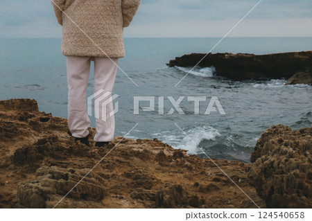 A lonely woman in a white coat stands on a stone seashore in the cold season and looks out to sea in the distance. Walking by the ocean in fall winter A lonely woman in a white coat stands on a stone seashore in the cold season and looks out to sea in the distance. Walking by the ocean in fall winter 124540658