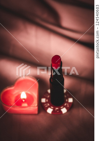 Red lipstick on a poker chip on a red satin background. Cosmetics product. Female beauty. Vogue fashion women lifestyle A burning heart-shaped candle. Red lipstick on a poker chip on a red satin background. Cosmetics product. Female beauty. Vogue fashion women lifestyle A burning heart-shaped candle. 124540683