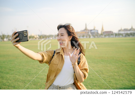Traveler asian woman in her 30s making a livestream or selfie on smartphone while explores Wat Phra Kaew emerald Buddha. Share the wonders of Thai heritage through her journey. 124542579