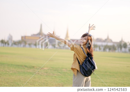 A Traveler Asian woman in 30s, bathed in golden glow of a Bangkok sunset, laughs with carefree joy, her arms outstretched as if to embrace freedom of the moment. Backpack strapped on. 124542583