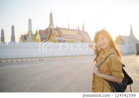 Traveler asian woman in her 30s, backpack slung over her shoulder, explores the intricate details of Wat Pra Kaew with childlike wonder. Sunlight dances on the golden rooftops. Traveler asian woman in her 30s, backpack slung over her shoulder, explores the intricate details of Wat Pra Kaew with childlike wonder. Sunlight dances on the golden rooftops. 124542599