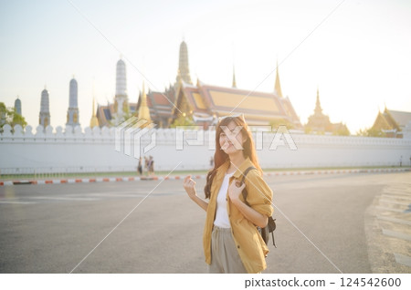 Traveler asian woman in her 30s, backpack slung over her shoulder, explores the intricate details of Wat Pra Kaew with childlike wonder. Sunlight dances on the golden rooftops. 124542600
