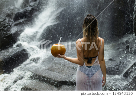 Beautiful sexy woman stands facing a waterfall in Bali and holds a coconut in her hand 124543130