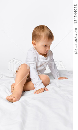 Focused little boy sitting on bed with curious look against white studio background. 124544028