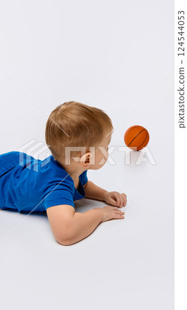 Toddler lying on belly with basketball roll away on floor against white background. 124544053