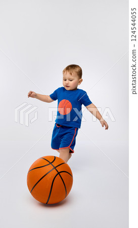 Boy playing with basketball, trying to kick, exploring physical skills against white background. 124544055