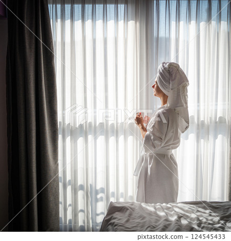 Beautiful young woman in a white bathrobe with a towel on her head stands near the window. 124545433