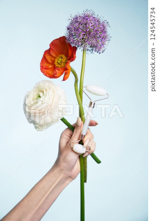 Hand holding flowers and a facial roller against a light background Hand holding flowers and a facial roller against a light background 124545734