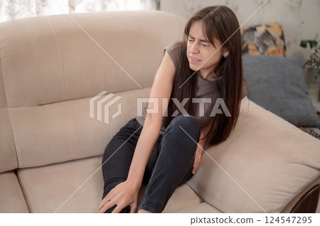 Young woman sitting on sofa experiencing discomfort in her knee while adjusting her posture in a cozy indoor setting Young woman sitting on sofa experiencing discomfort in her knee while adjusting her posture in a cozy indoor setting 124547295