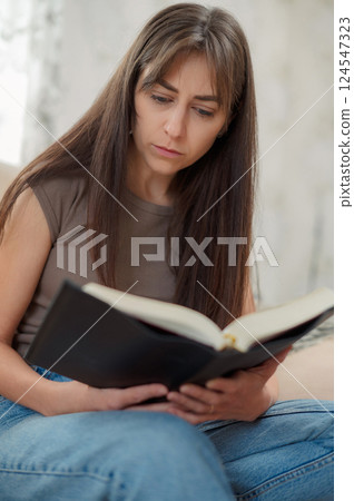Young woman engrossed in a captivating book, sitting comfortably on a sofa in the cozy atmosphere of her living room 124547323