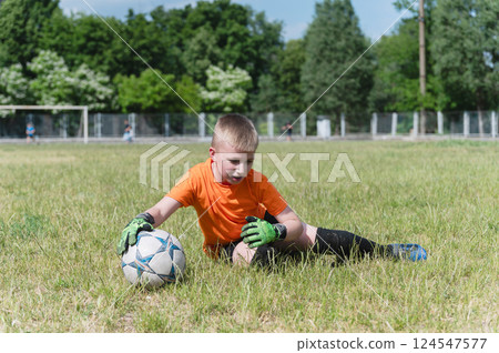 Goalkeeper boy sitting on grass holding soccer ball. 124547577