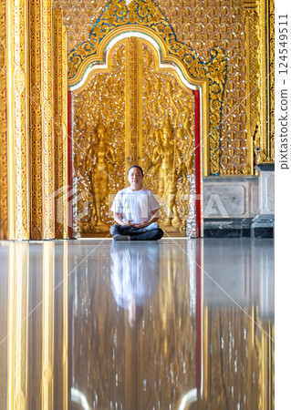 Asian Man meditates on the ground with reflection in the sunlight pouring into the temple church with gold gold Thai pattern blur background. 124549511