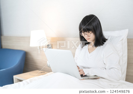 Glasses Asian cute woman works and plays laptop notebook on the bed in Work From Home period. 124549876