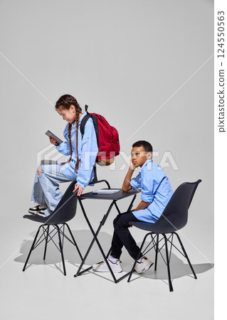 Girl reads tablet at desk while boy looks thoughtfully, representing different approaches to study on knowledge day. Education concept. 124550563