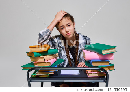 Young girl, student, sits focused at desk surrounded by colorful books, pen held thoughtfully while studying during lesson time, with exhausted expression against plain background. Young girl, student, sits focused at desk surrounded by colorful books, pen held thoughtfully while studying during lesson time, with exhausted expression against plain background. 124550595