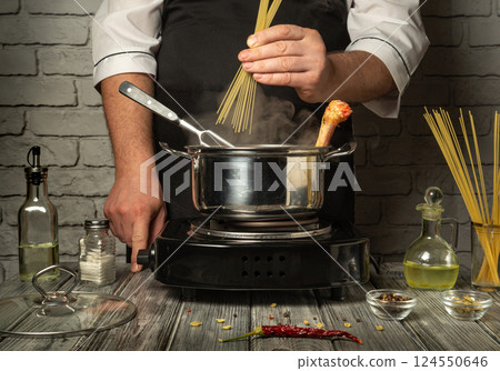 A chef prepares to cook pasta by adding dry spaghetti to boiling water in a metal pot. Surrounding ingredients include olive oil, spices, and herbs on a rustic table A chef prepares to cook pasta by adding dry spaghetti to boiling water in a metal pot. Surrounding ingredients include olive oil, spices, and herbs on a rustic table 124550646