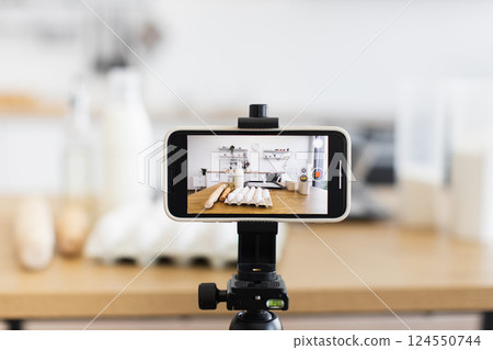 Smartphone on tripod recording video in modern kitchen with baking ingredients and blurred background. Captures process of preparing food, ideal for tutorials, cooking videos, or blogging Smartphone on tripod recording video in modern kitchen with baking ingredients and blurred background. Captures process of preparing food, ideal for tutorials, cooking videos, or blogging 124550744