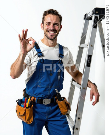 Happy male worker in blue overalls giving okay gesture beside ladder 124551673