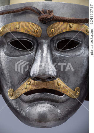 Historical metallic combat helmet with integrated face mask. Pointed steel helmet featuring a detailed metallic face mask with eye openings, decorative elements, and side protection flaps. 124553757