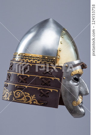 Historical metallic combat helmet with integrated face mask. Pointed steel helmet featuring a detailed metallic face mask with eye openings, decorative elements, and side protection flaps. 124553758