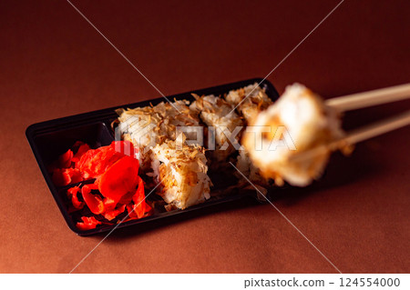 Chopsticks taking portion of sushi roll. Sushi rolls with salmon cream cheese and cucumber in take away package. Food delivery concept. 124554000