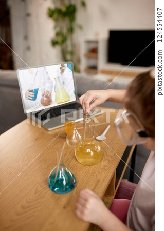Girl carefully mixing liquids in beaker while following virtual science class on laptop screen. Online chemistry class. Girl carefully mixing liquids in beaker while following virtual science class on laptop screen. Online chemistry class. 124554407
