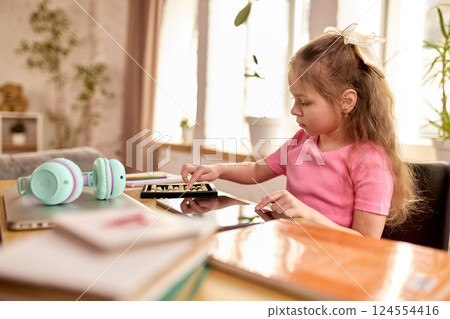 Girl in pink shirt practicing counting on abacus next to tablet during quiet online math class in cozy home environment. Girl in pink shirt practicing counting on abacus next to tablet during quiet online math class in cozy home environment. 124554416