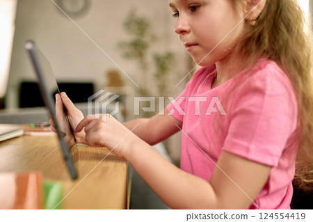 Focused girl in pink shirt interacting with tablet during remote learning session at home, using touchscreen attentively. Focused girl in pink shirt interacting with tablet during remote learning session at home, using touchscreen attentively. 124554419