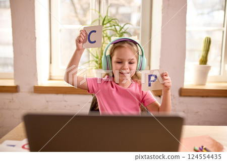 Smiling schoolgirl showing flashcards with letters C and P to laptop with teacher during fun alphabet lesson at home 124554435