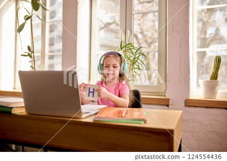 Girl, elementary school pupil in headphones holding flashcard with letter H while learning online at home with study materials around. 124554436
