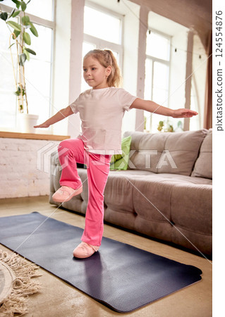 Little girl balancing on one leg during online workout session in living room on blue fitness mat in activewear. 124554876