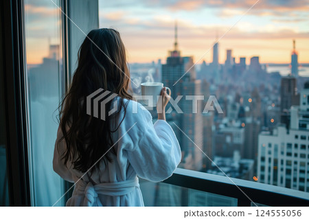 Back view of a woman standing on a city balcony, holding a morning coffee mug while wearing a fluffy white robe. Captures peaceful moments of relaxation in an urban setting during sunrise. 124555056
