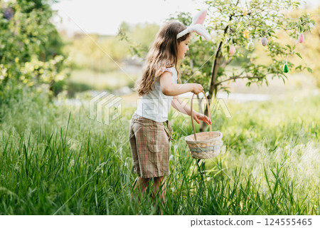 Easter egg hunt. Girl child Wearing Bunny Ears Running To Pick Up Egg In Garden. Easter tradition. Baby with basket full of colorful eggs. Easter egg hunt. Girl child Wearing Bunny Ears Running To Pick Up Egg In Garden. Easter tradition. Baby with basket full of colorful eggs. 124555465