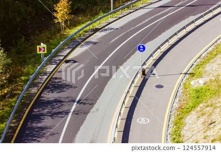 Top view of the paved road in the corner in the forest. Carriageway and sidewalk for pedestrians and bicycle lane with markings. Road signs. Safety. 124557914