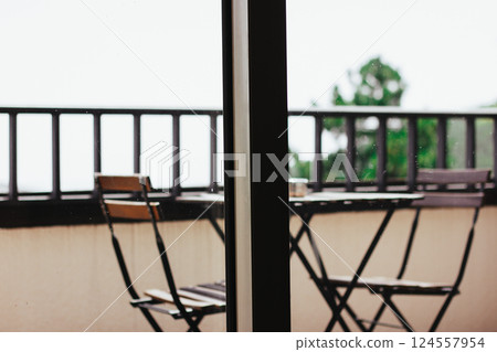 Wooden table and chairs on balcony behind a panoramic window. Garden furniture at home. Empty patio in a house in a forest. Railing on terrace. 124557954