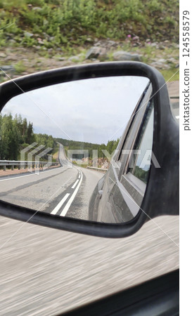 A winding rural road surrounded by lush green forest is captured through the side mirror of a moving car, symbolizing travel, freedom, and exploration. The overcast sky adds a moody tone to this 124558579