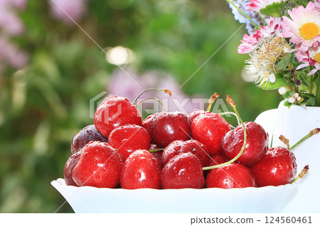 A cup of cherries on a sunny table in a flowered garden, selective focus. Sweet cherry with droplets and a branch of lilac in the background in the garden, the effect of natural bokeh in the A cup of cherries on a sunny table in a flowered garden, selective focus. Sweet cherry with droplets and a branch of lilac in the background in the garden, the effect of natural bokeh in the 124560461