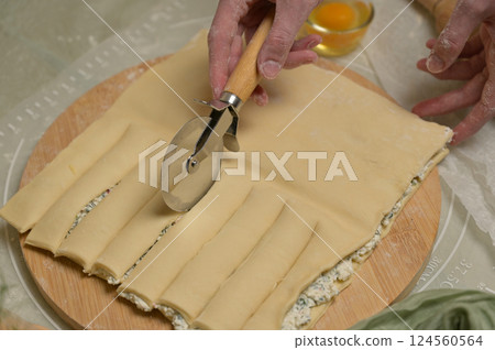 Close up square sheet of dough cut into strips with woman's hands on wooden board. Cutter, egg on kitchen table. Home cooking baking puffs Close up square sheet of dough cut into strips with woman's hands on wooden board. Cutter, egg on kitchen table. Home cooking baking puffs 124560564